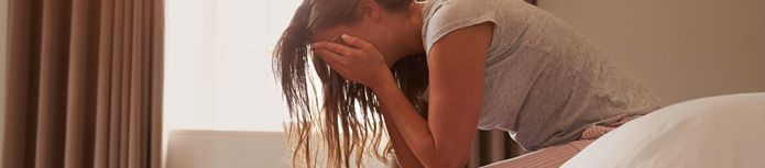 banner: a close shot of a woman who sits at the edge of her bed and covers her face with her two hands