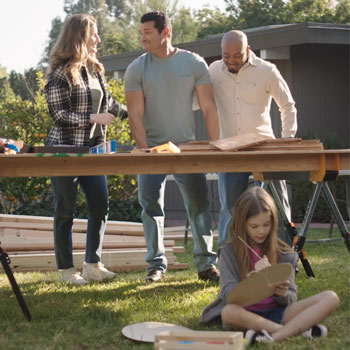 Three adults stand at a table with wood and tools, while a young girl sits on the grass painting a wooden craft in a backyard setting.